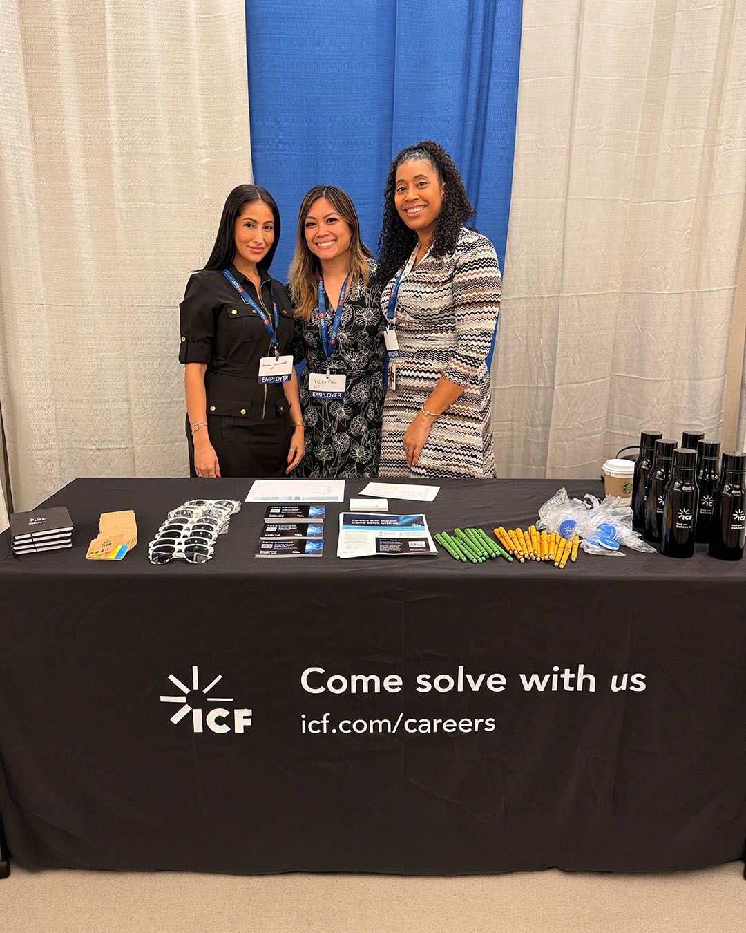 Three employees stand behind an ICF booth table with brochures, sunglasses, pens, and branded bottles, with a banner reading “Come solve with us” and icf.com/careers