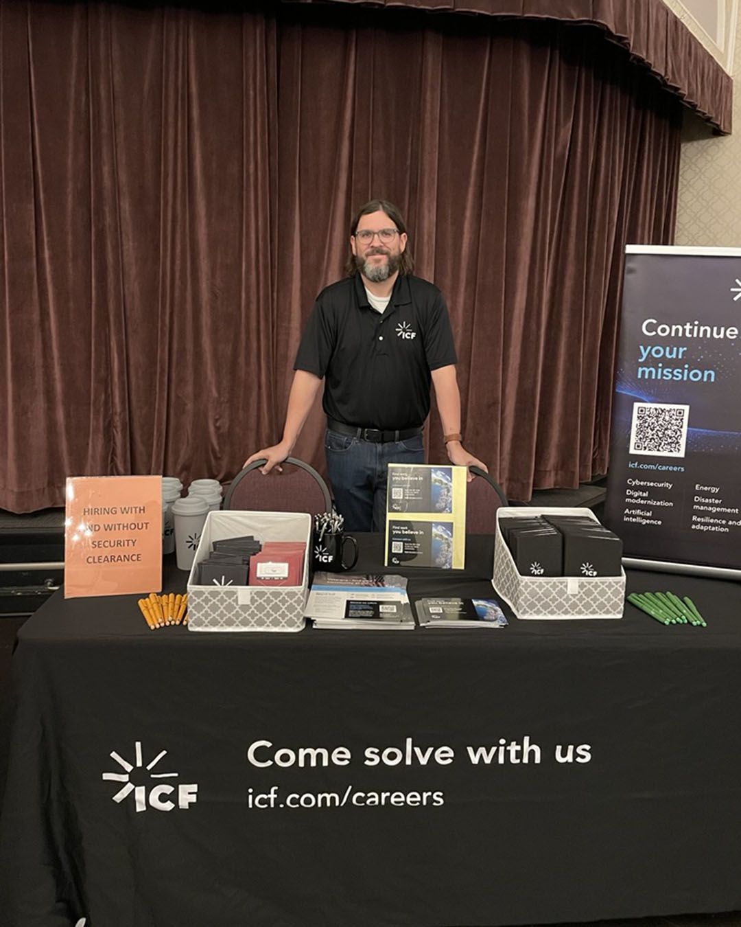 An ICF employee at a job fair standing in front of a table cloth that has a message printed on it that reads:Come solve with us. icf.com/careers