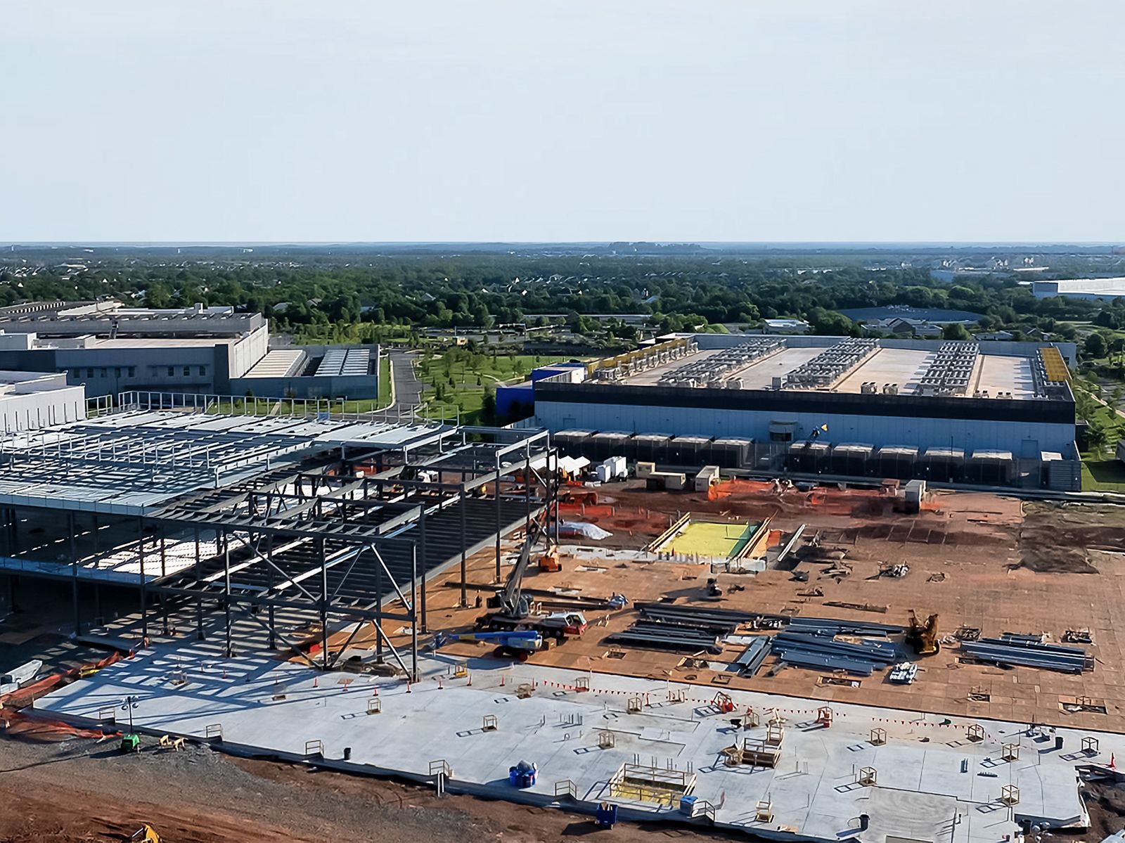 An aerial view of a data center build in progress in front of a horizon of trees and blue sky.