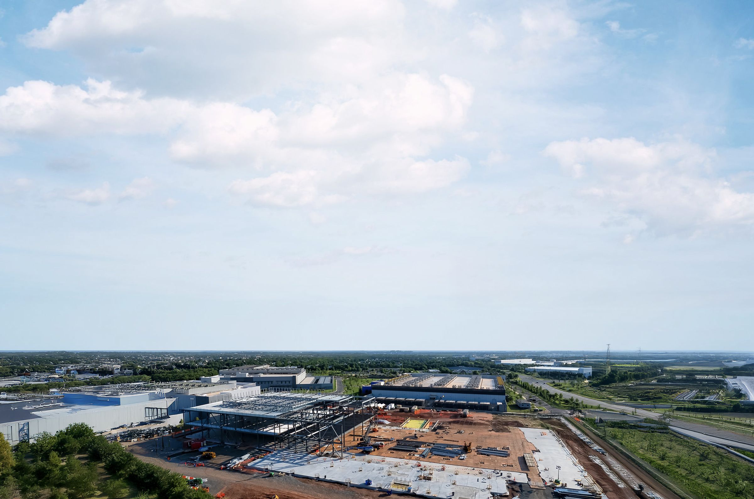 An aerial view of a data center build in progress in front of a horizon of trees and blue sky.