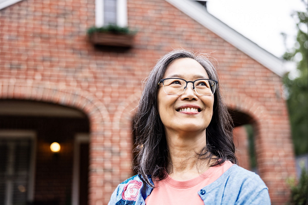 A woman with glasses and shoulder length hair smiles in front of a red brick house.